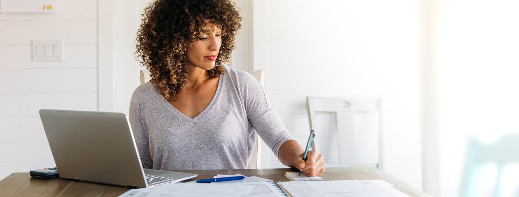 A woman jots down some notes next to her laptap.