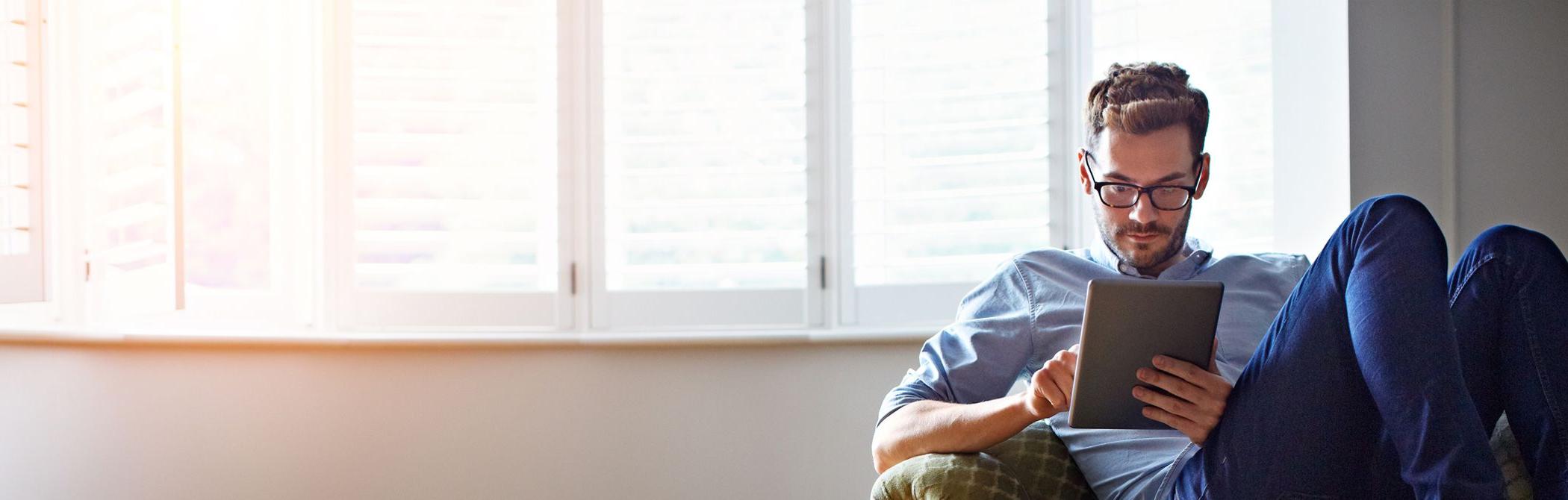 A Millennial reads his tablet in front of some windows.